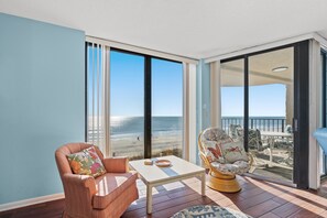 A room with a view of the beach, a chair, a table, and a wicker chair.