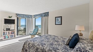 Un cuarto de hotel con una cama grande, un televisor, una estantería, una silla junto a la ventana y vista al océano.