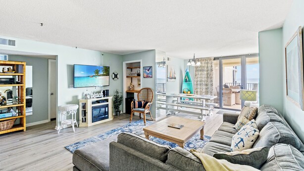 A spacious living room with a large sofa, a coffee table, and a television. There is a balcony with outdoor furniture visible through the glass doors. - Sea Castle 2F-Oceanfront 3 Bedroom- Pool & Hot Tub (North Myrtle Beach)