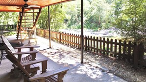 A covered outdoor seating area with wooden benches and a ladder, overlooking a natural landscape with trees and a wooden fence.