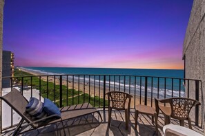 A balcony with a view of the beach and ocean, equipped with chairs and a table.