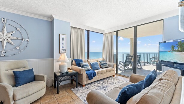 Een woonkamer met een bank, fauteuils, een televisie en een groot raam met uitzicht op zee.