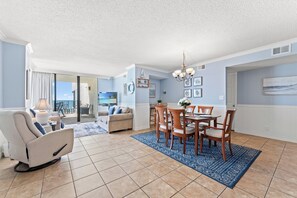 A modern living room with a sofa, dining table, and chairs, a balcony with a view, and a kitchen area.