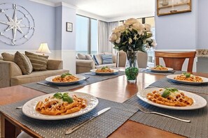 A dining area with a table set for four, featuring plates of pasta, a vase with flowers, and a compass decoration on the wall.