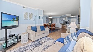 Een woonkamer met een televisie, een beige bank, blauwe kussens, een salontafel en een eethoek met stoelen en een tafel.
