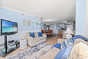 Una sala de estar con televisión, sofá color beige, cojines azules, una mesa de centro y una zona de comedor con sillas y una mesa.