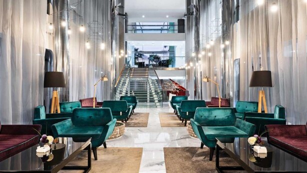 A modern hotel lobby with teal upholstered chairs, a central staircase, and a glass railing overlooking the interior.