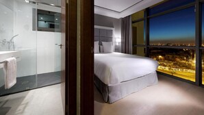 A modern hotel room with a large bed, a glass-enclosed shower, and a city view through large windows.