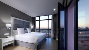 A modern hotel room with a large bed, a nightstand, and a view of the cityscape through large windows.