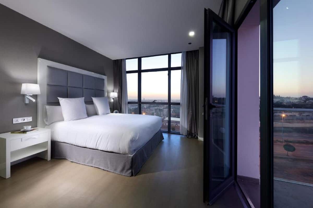 A modern hotel room with a large bed, a nightstand, and a view of the cityscape through large windows.