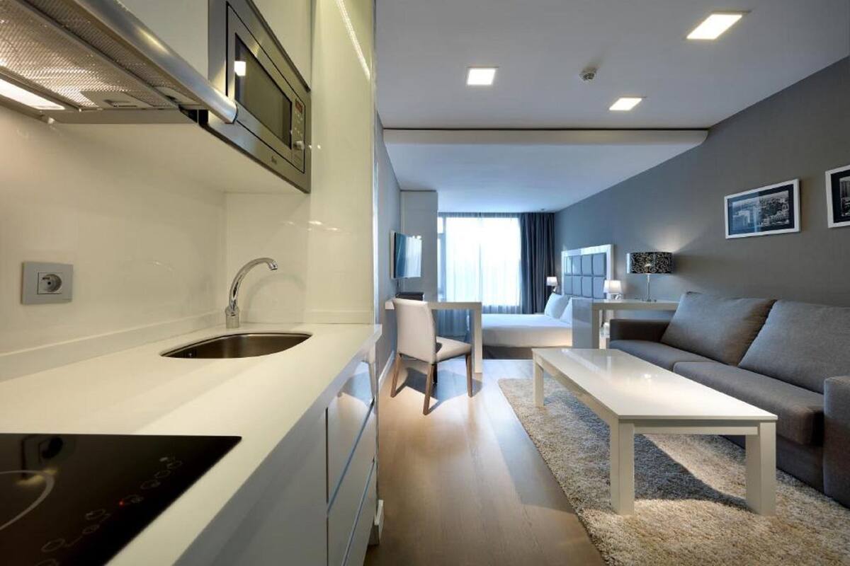 A modern kitchen with a built-in stove, a sink, and a microwave. There is a dining area with a white table and a sofa. The room has a neutral color scheme and a large window providing natural light.