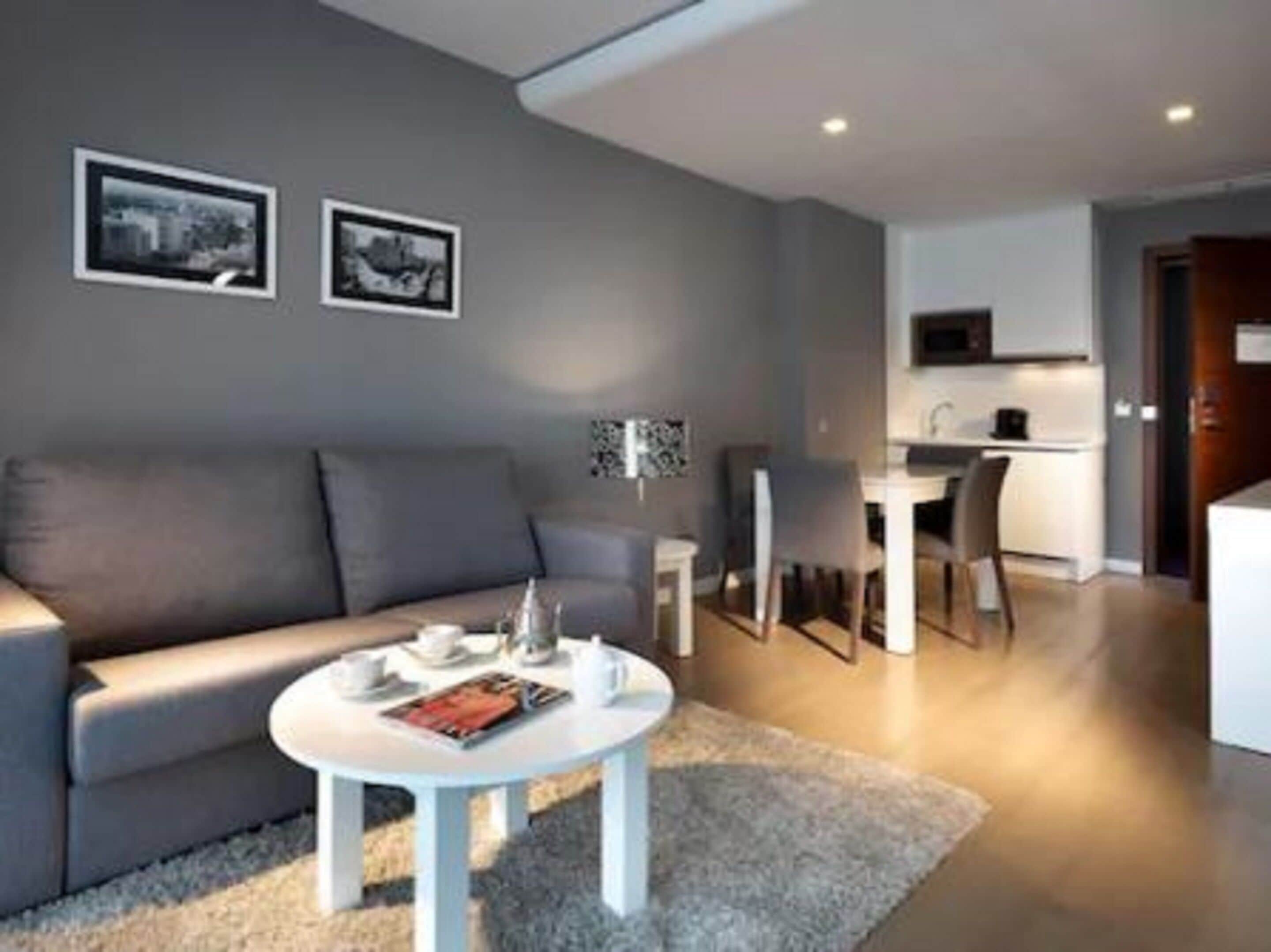 Un salon moderne avec un canapé gris, une table basse ronde et une petite table blanche. Deux tableaux encadrés sont accrochés au mur, une lampe est posée sur la table basse, et on aperçoit un coin cuisine au fond de la pièce.