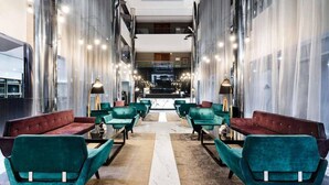 A modern hotel lobby with teal and maroon seating, a central marble pathway, and a view of the lobby area through the glass walls.