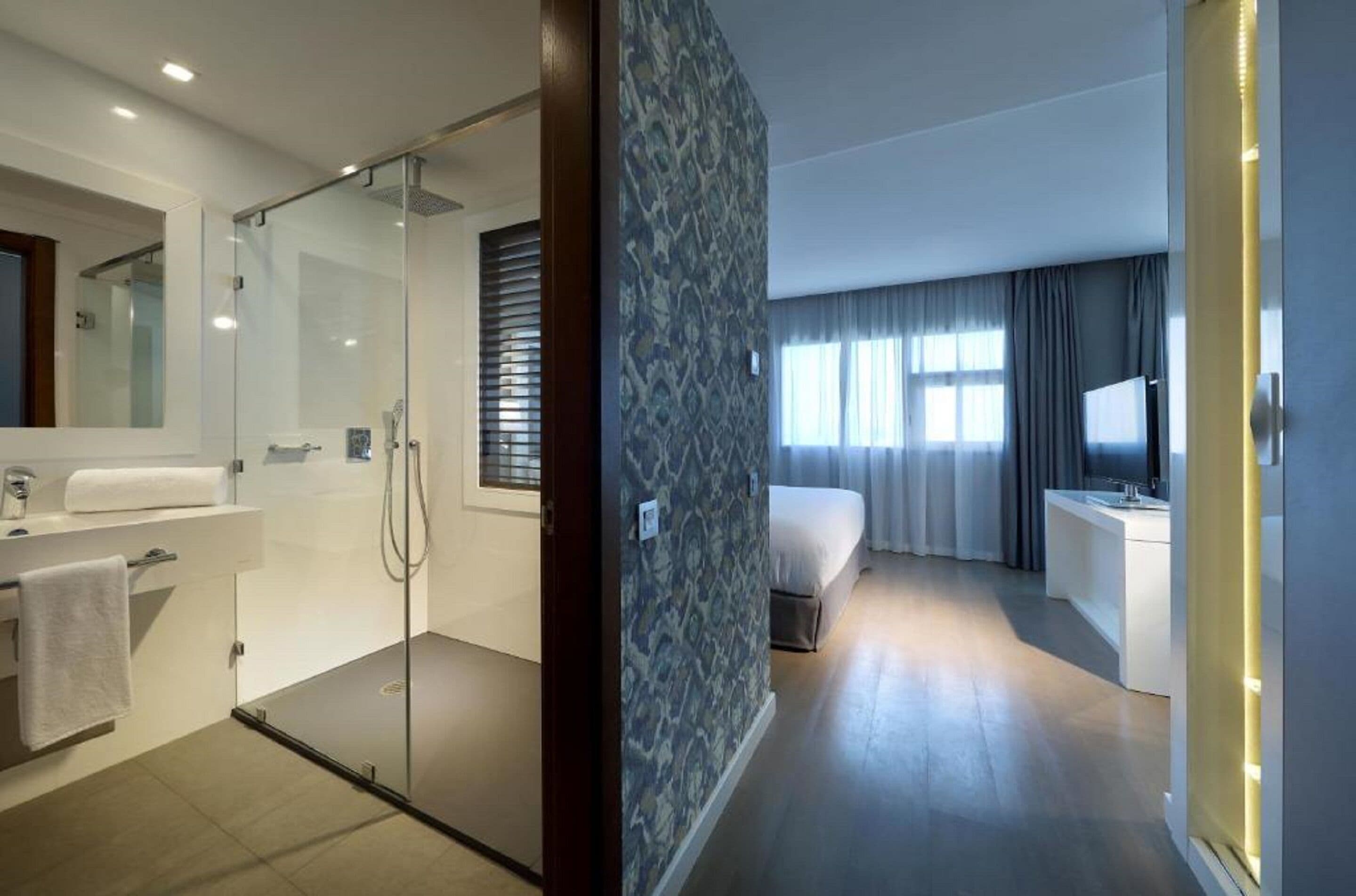 Una habitación de hotel moderna con ducha envuelta en vidrio, baño independiente, cama y una pequeña zona de estar.