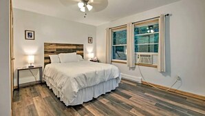 Een slaapkamer met een houten hoofdbord, een bed met wit beddengoed, een nachtkastje met een lamp, een plafondventilator en een raam met gordijnen.