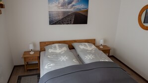 Un dormitorio con una cama, dos mesitas de noche, una lámpara de noche, una alfombra y un cuadro colgado en la pared que muestra una escena costera.