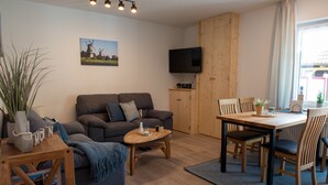 Una sala de estar moderna con un sofá, una mesa de centro y una mesa de comedor. Hay un televisor montado en la pared y una ventana con cortinas.