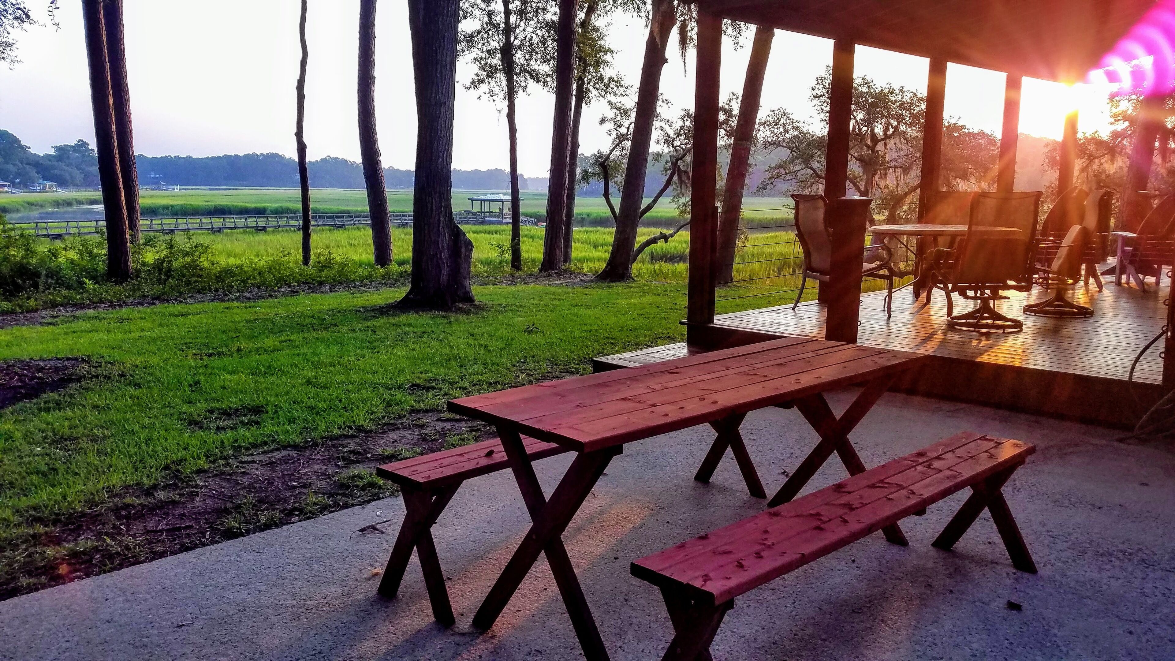 An outdoor patio with a wooden table and benches, overlooking a grassy field and trees.