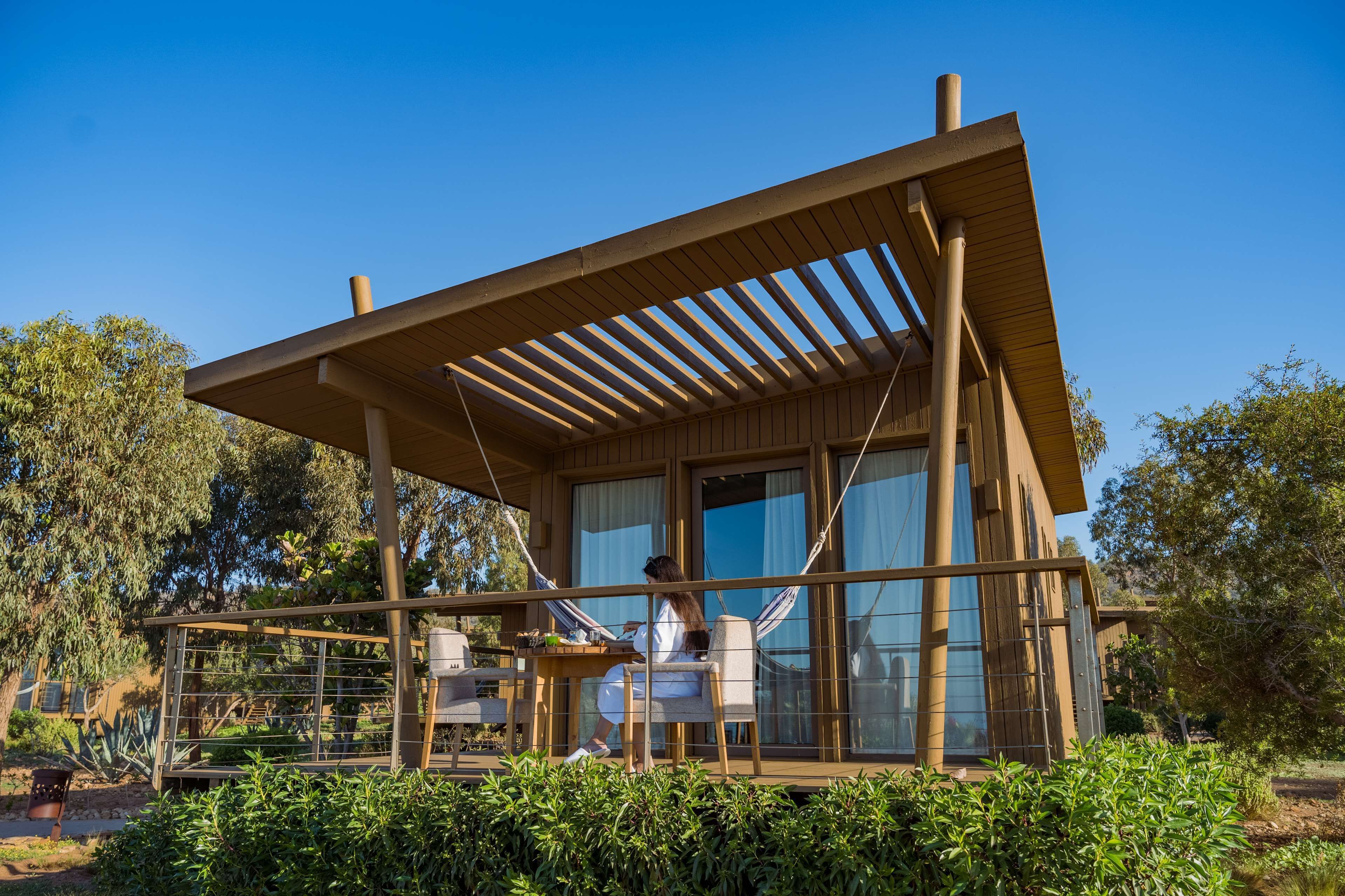 Una cabaña de madera moderna con un porche cubierto, ventanas amplias y vistas a la vegetación circundante.