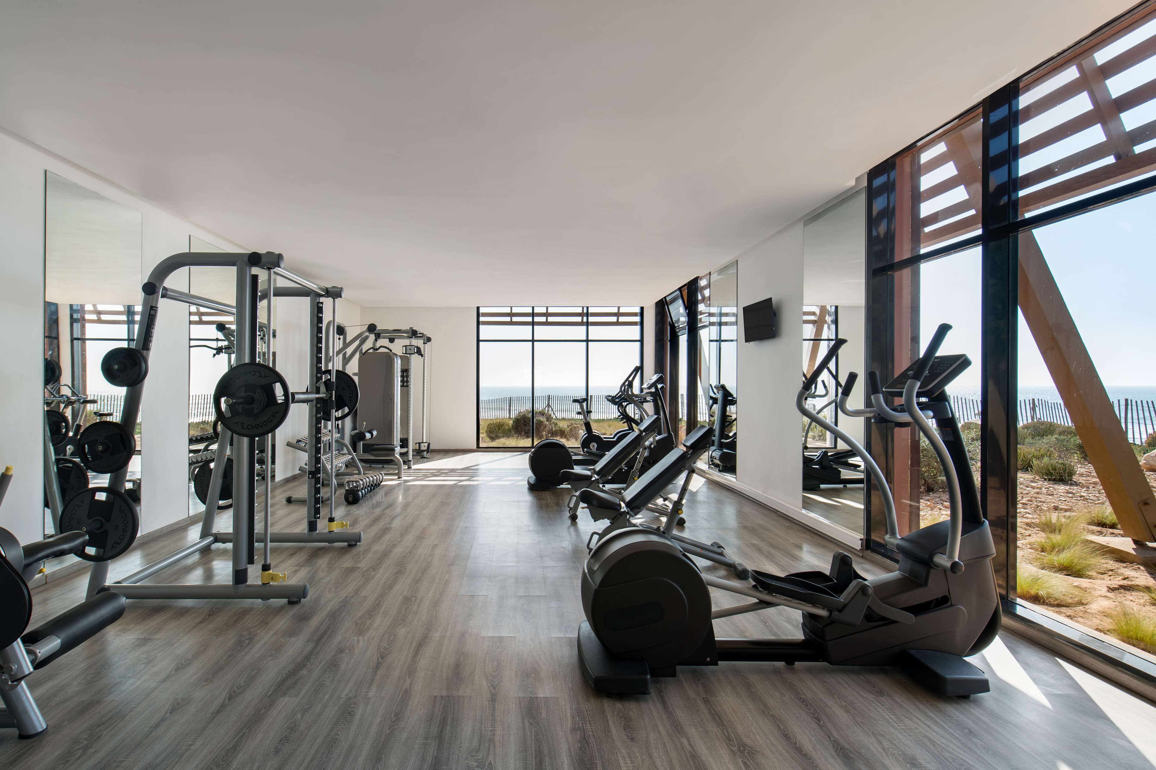 A modern gym with various exercise equipment, large windows, and a wooden floor.