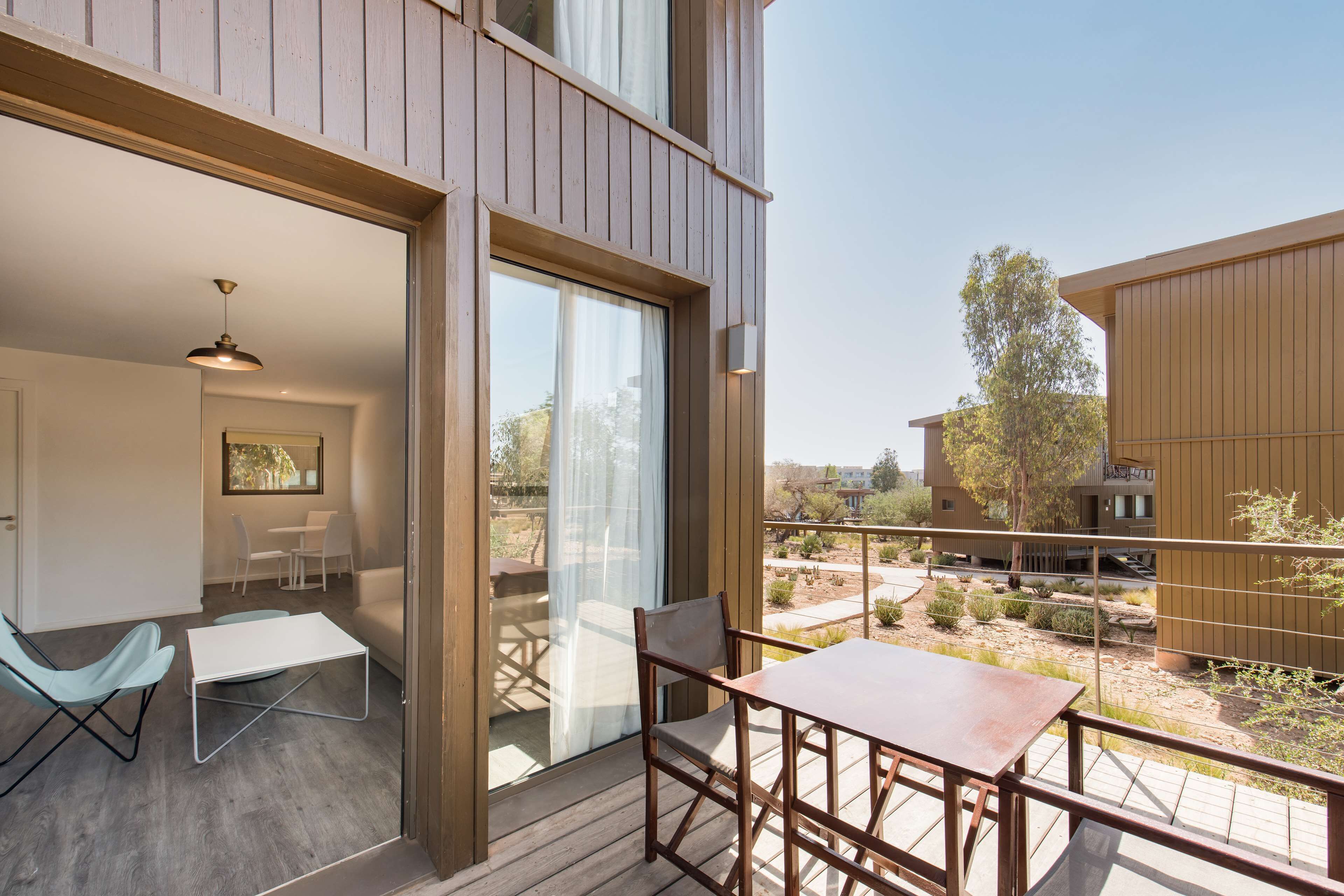 Ein moderner Balkon mit Holztable und -stühlen, eine Glastür zu einer Terrassenfläche und Blick auf eine Wohngegend.