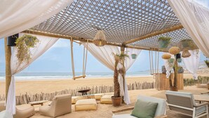 A beachside outdoor seating area with cushioned chairs and tables, a wooden deck, and hanging planters. - Radisson Blu Resort, Taghazout Bay Surf Village (Taghazout)