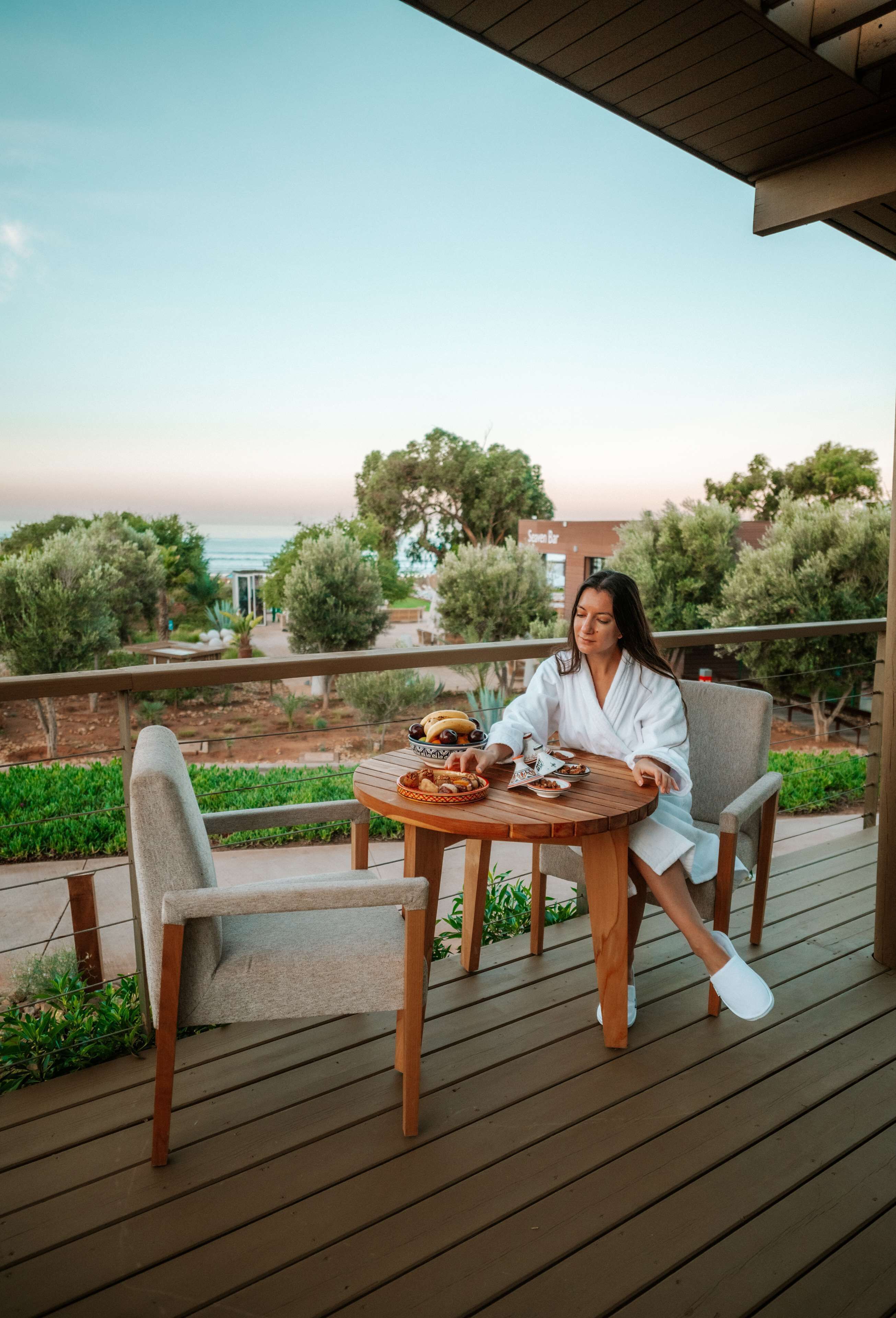 A woman sitting at a wooden table on a deck with a view of the ocean and trees.