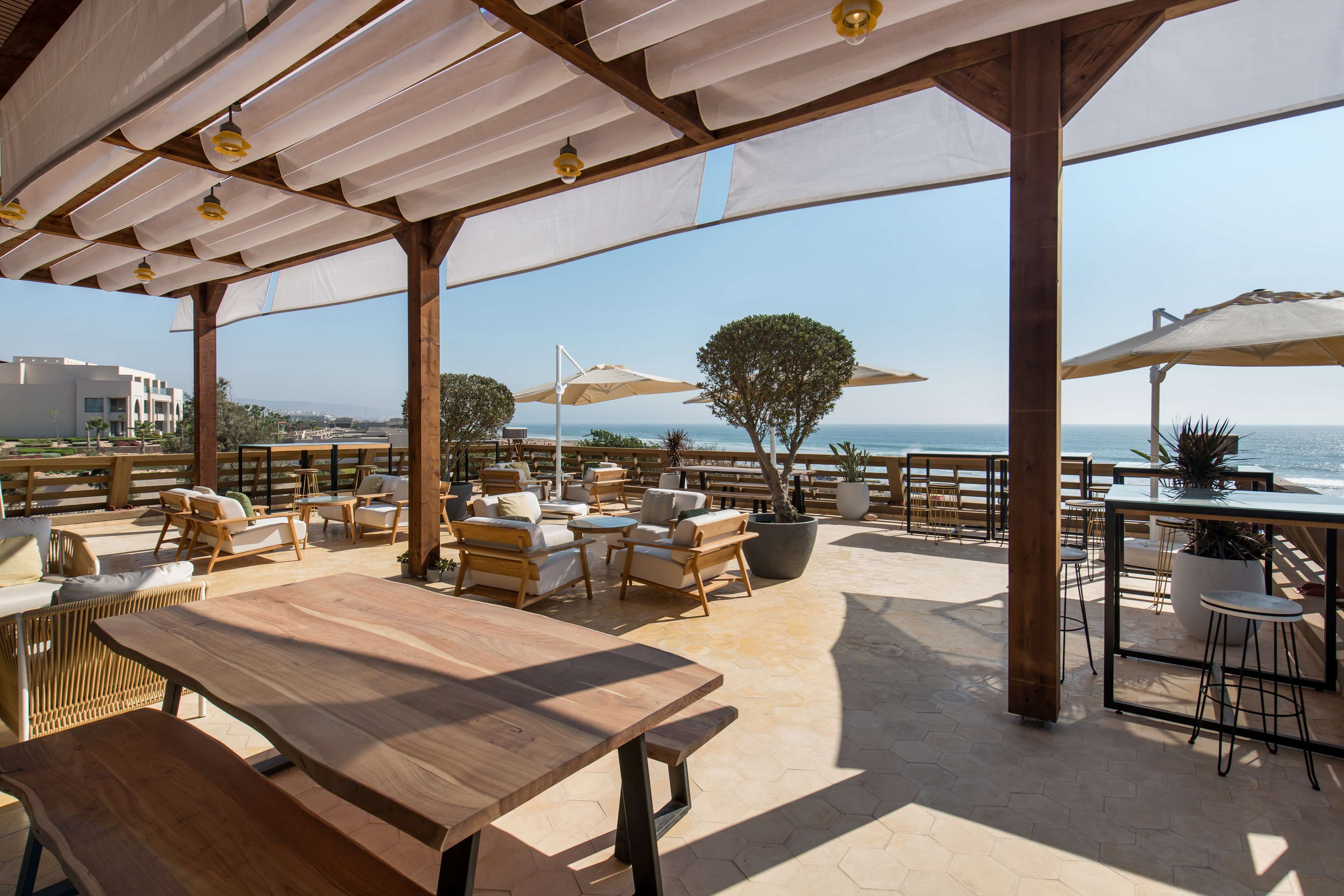 Eine Terrassengaststätte mit Holztischen und -stühlen, Sonnenschirmen und Blick auf das Meer.