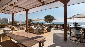 Eine Terrassengaststätte mit Holztischen und -stühlen, Sonnenschirmen und Blick auf das Meer.