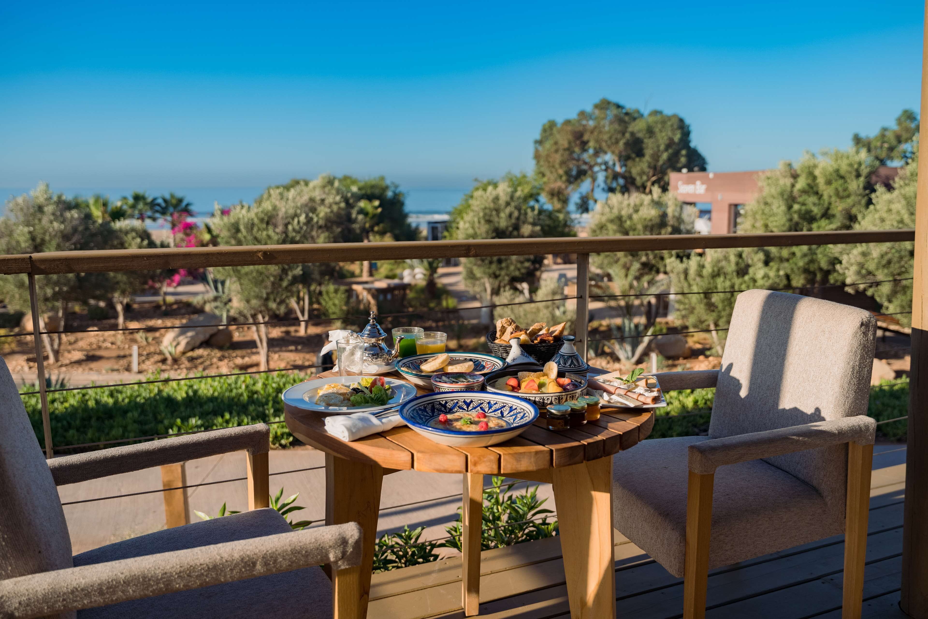 Una terraza de madera con una mesa dispuesta para una comida, con vistas a un paisaje pintoresco con árboles y vista al mar.