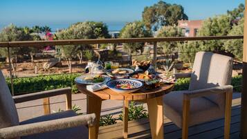 Une terrasse en bois avec une table dressée pour un repas, offrant une vue panoramique sur un paysage verdoyant, avec des arbres et la mer à l’horizon.