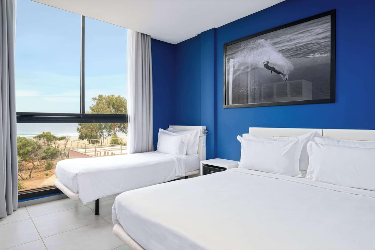 A hotel room with two beds, a large window with a view of the beach, and a framed picture on the wall.