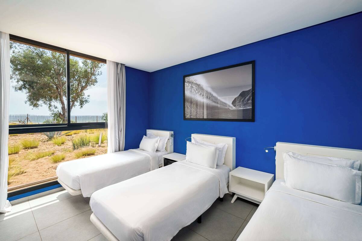 A hotel room with two beds, a large window with a view of a beach and trees, and a framed picture on the wall.
