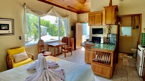 A room with a bed, a dining table, chairs, a kitchen area with a microwave, and a large window offering a view of greenery outside. - Belizean Reef Suites (San Pedro)