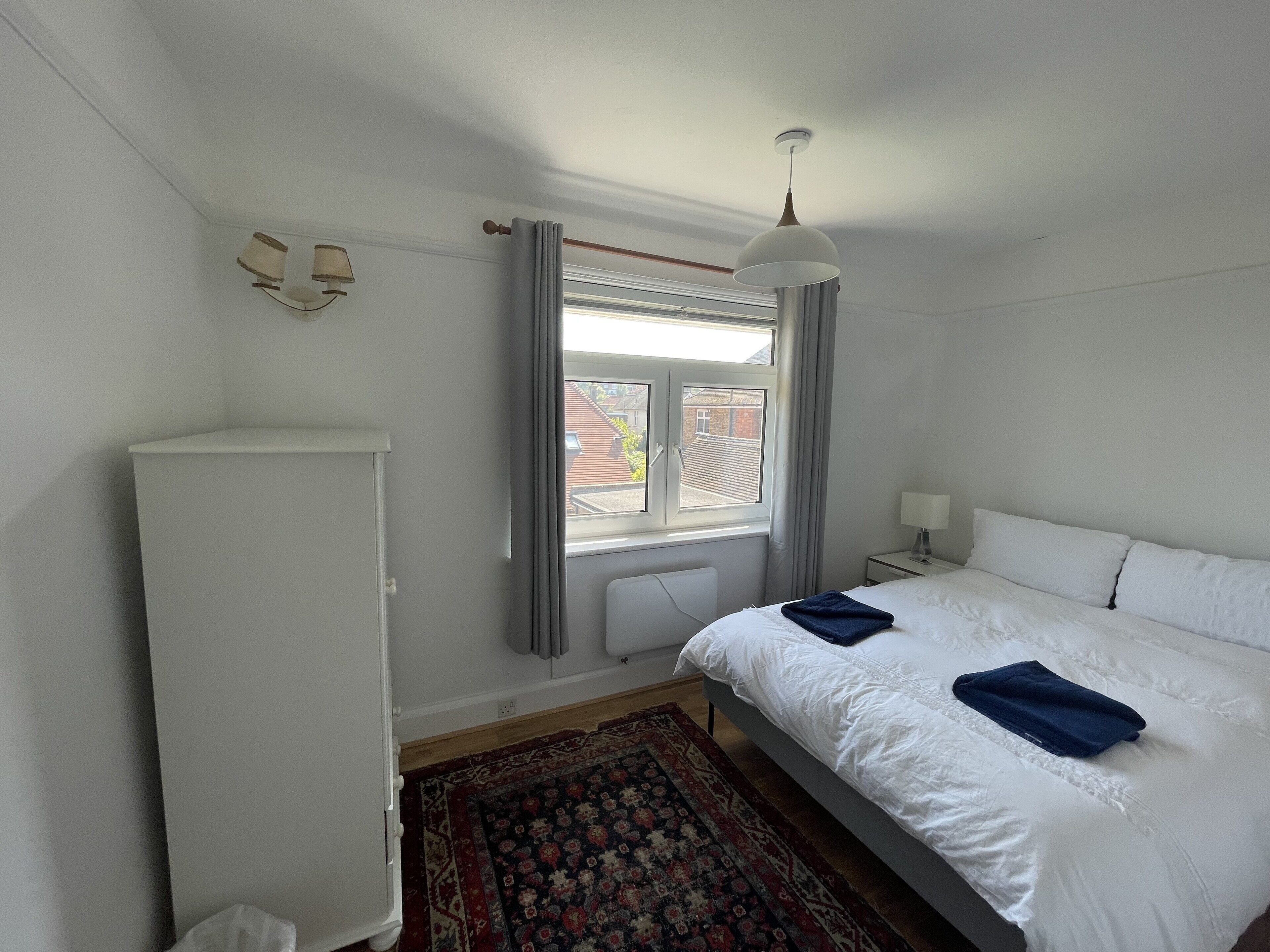 Un dormitorio con cama, radiador, ventana con cortinas, mesita de noche con lámpara y armario.