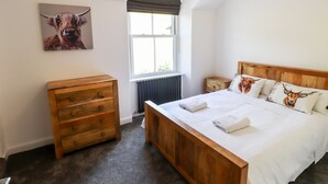 Une chambre avec un lit en bois, une commode en bois, un radiateur, une fenêtre avec des stores et un tableau représentant une vache accroché au mur.