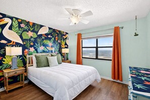 A bedroom with a bed, a ceiling fan, a window with curtains, and a wall with a tropical mural.