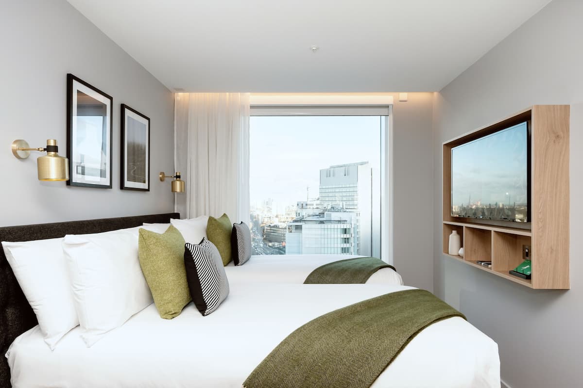 Une chambre d’hôtel moderne avec un grand lit, un téléviseur fixé au mur et une vue sur la ville par la fenêtre.