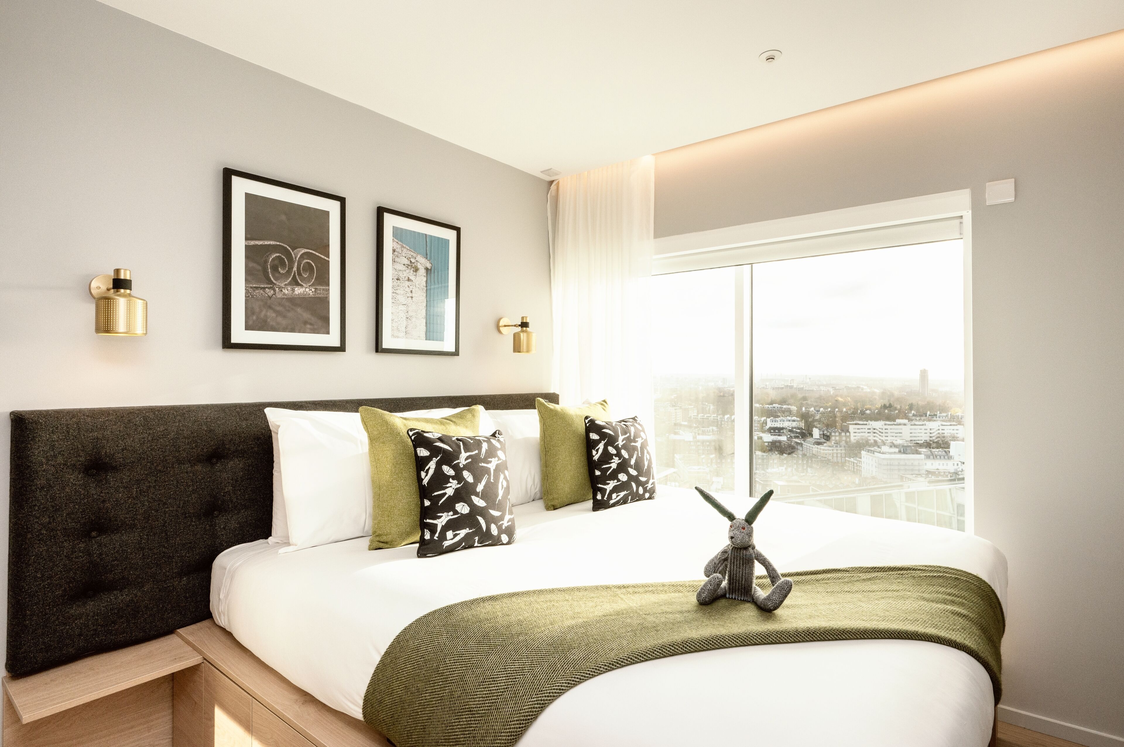A modern hotel room with a large bed, a view of the city, and two framed pictures on the wall.
