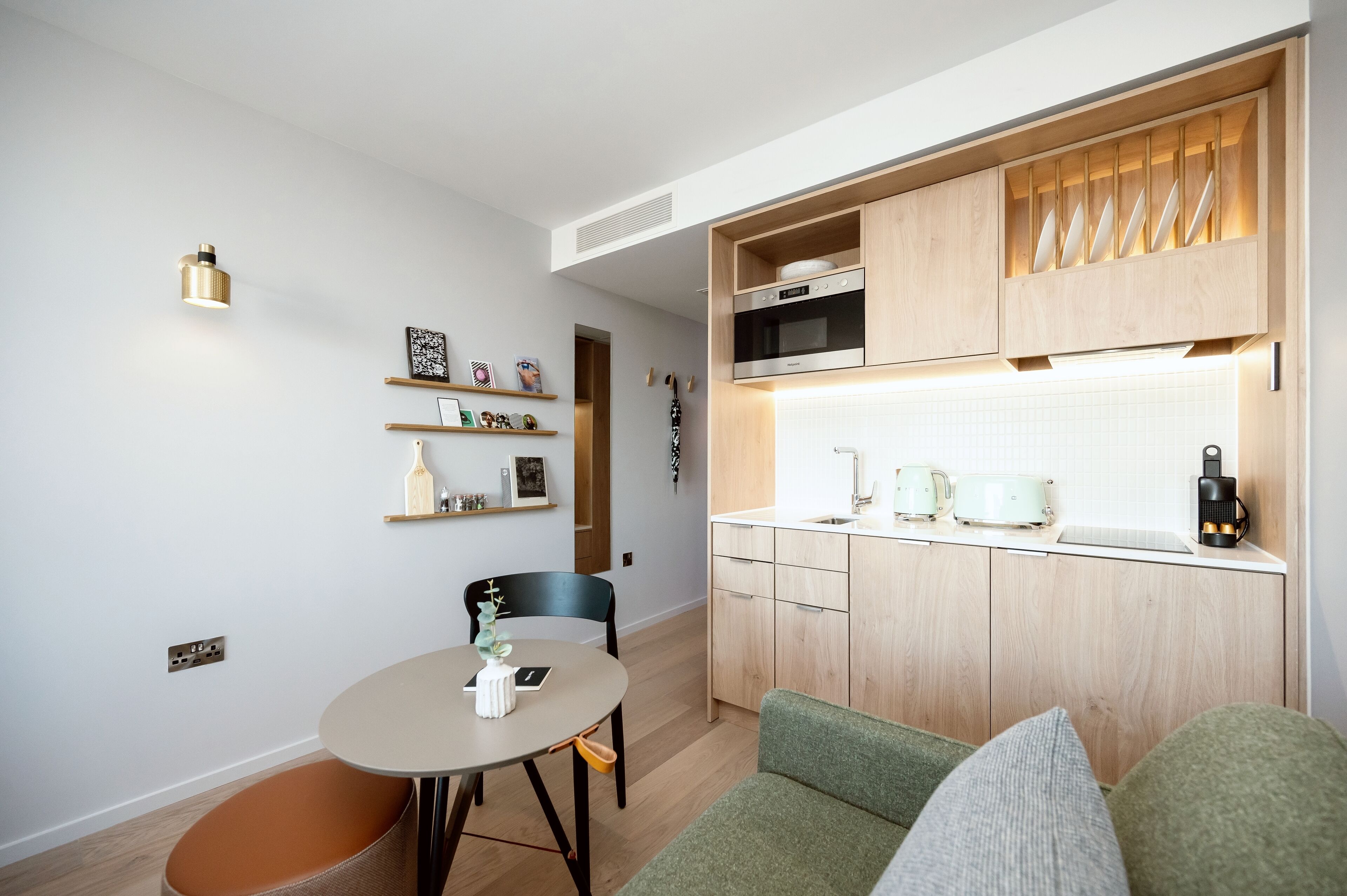 Une cuisine compacte dotée de meubles en bois, d’un four à micro-ondes et d’une bouilloire. On y trouve également un coin repas avec une table et des chaises, ainsi qu’une petite étagère où sont disposés des objets décoratifs.