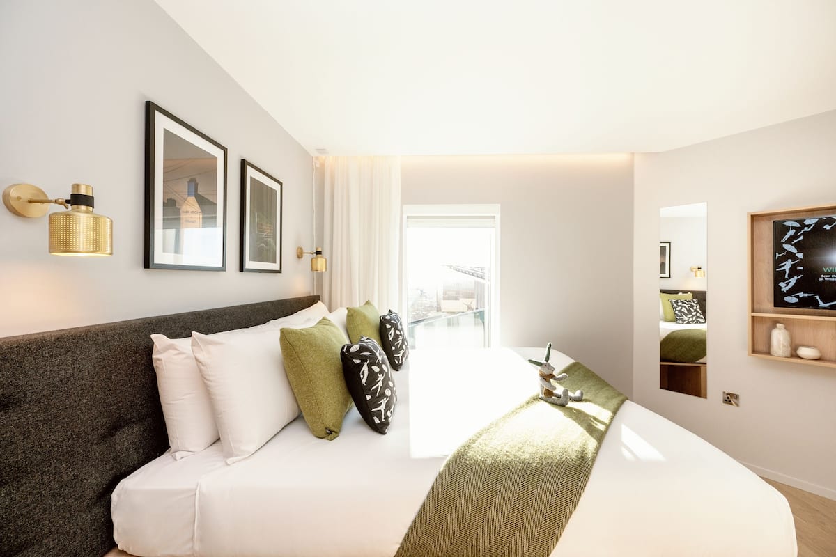Une chambre d’hôtel moderne avec un grand lit, une tête de lit sombre et une vue sur la ville par la fenêtre.