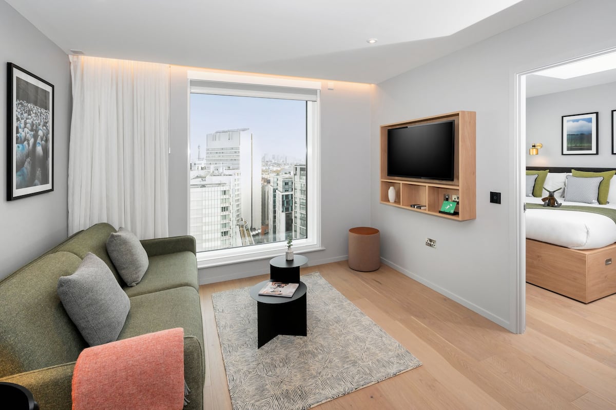 Un appartement moderne doté d’un canapé vert, d’un téléviseur à écran plat, d’un lit avec un cadre en bois et d’une grande fenêtre offrant une vue sur la ville.