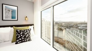 A hotel room with a large window offering a city view, a neatly made bed with white and green pillows, and a framed picture on the wall. - Wilde Aparthotels, London, Paddington (London)