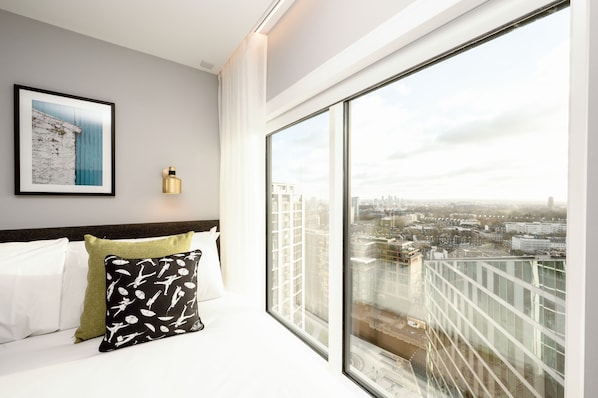 A hotel room with a large window offering a city view, a neatly made bed with white and green pillows, and a framed picture on the wall.
