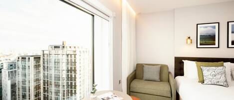 A modern hotel room with a large window, a bed, a sofa, a small table, and two framed pictures on the wall.