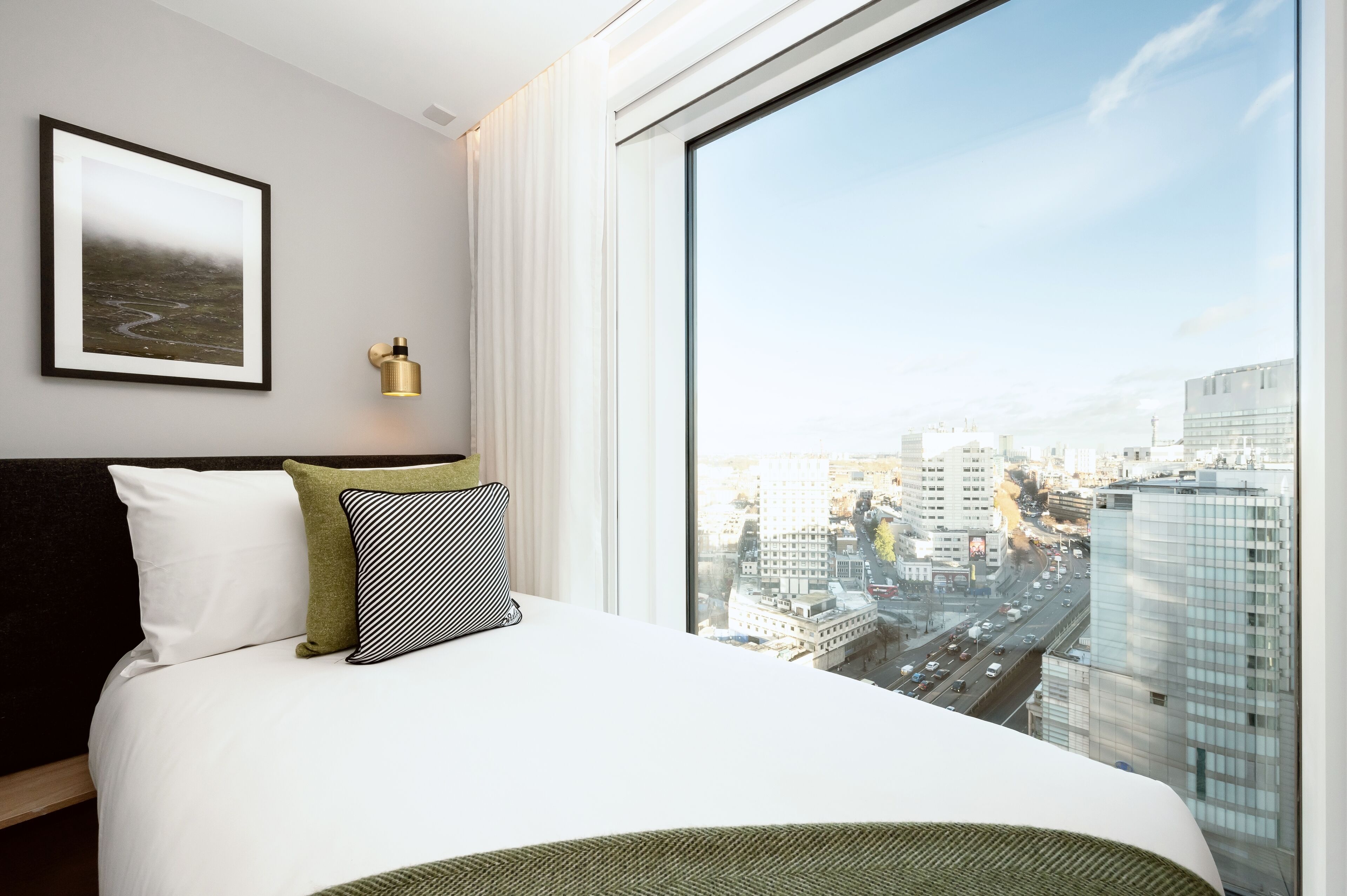 A modern hotel room with a large window offering a city view, a neatly made bed with pillows, and a framed picture on the wall.