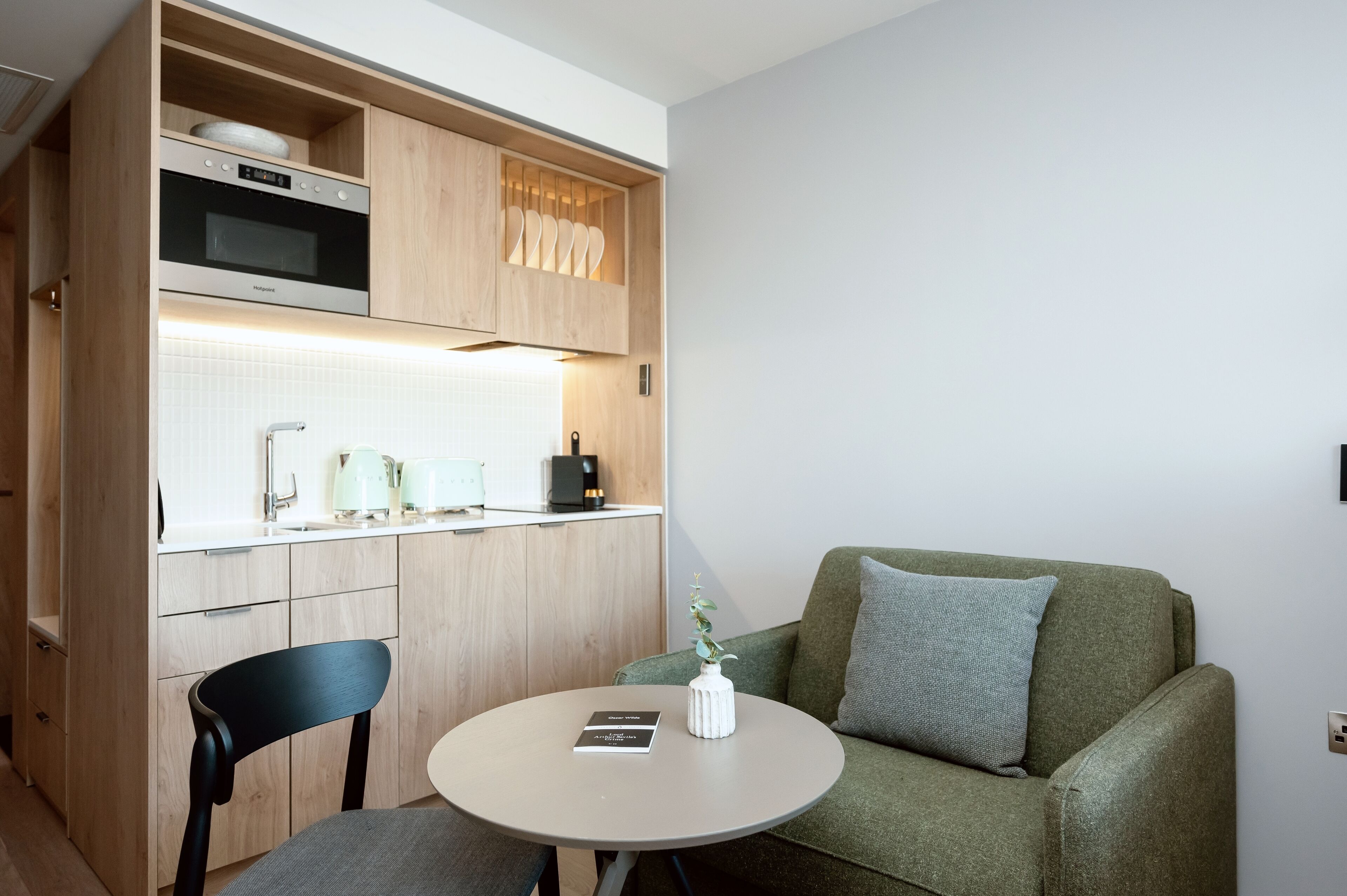 A compact kitchen with wooden cabinets, a microwave, and a coffee maker. There's a round table with a book and a vase with a plant. A green sofa with a pillow is against the wall.