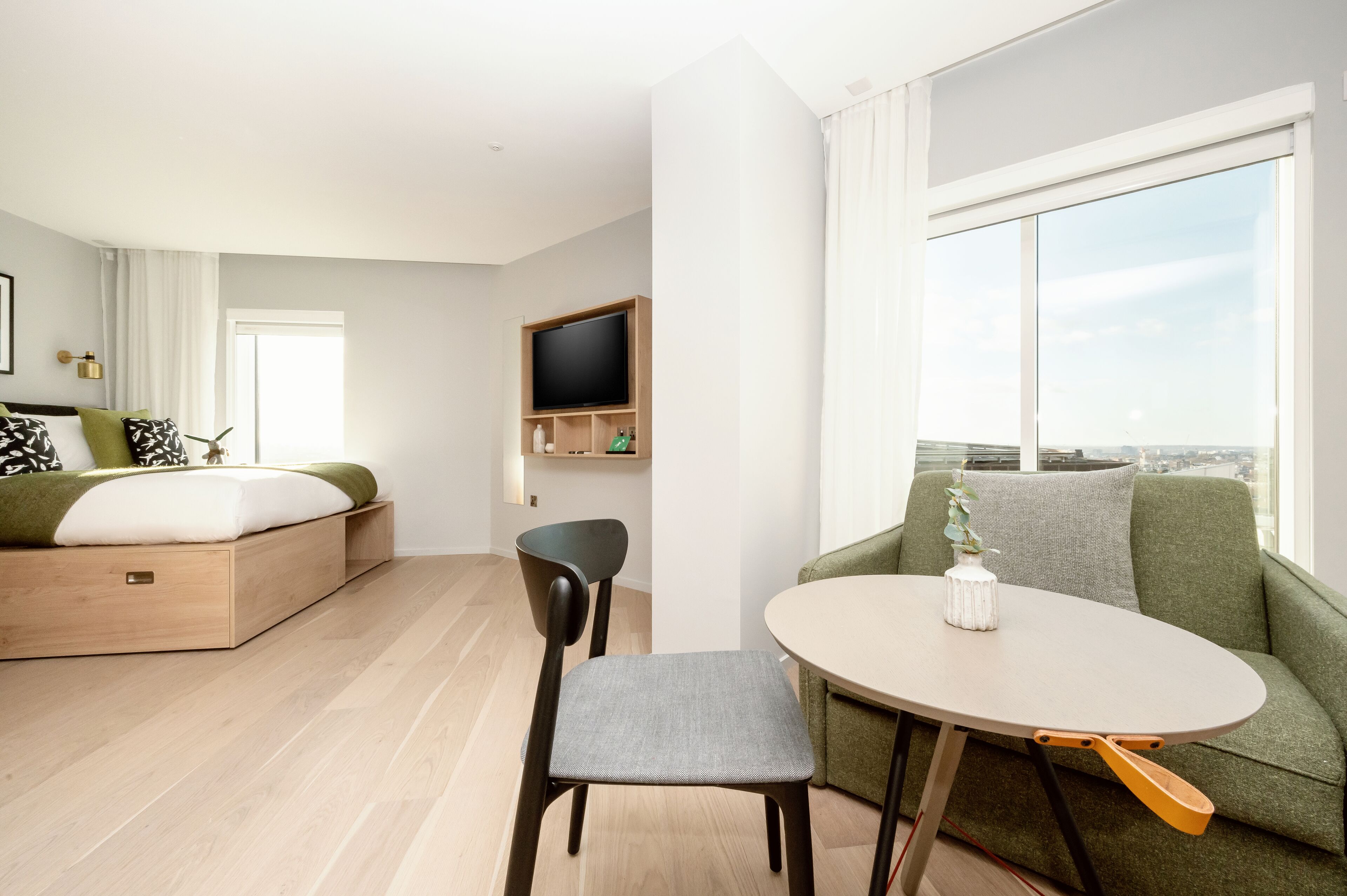 Una habitació d'hotel moderna amb llit, zona de menjador amb taula i cadires, televisió de pantalla plana i una finestra gran amb vista a la ciutat.