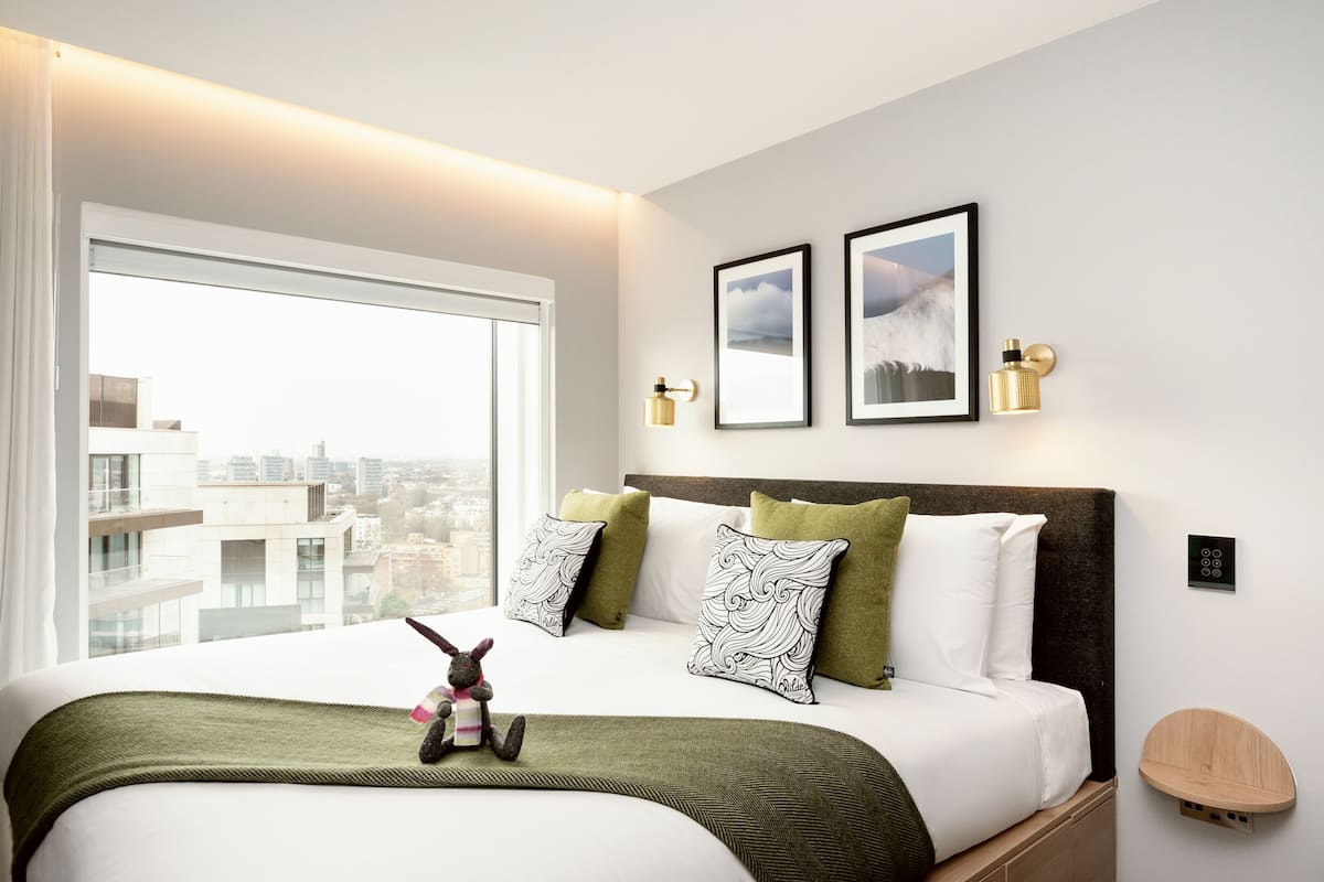 Une chambre d’hôtel moderne avec un grand lit, une vue sur la ville, des œuvres d’art encadrées et une petite table de chevet.