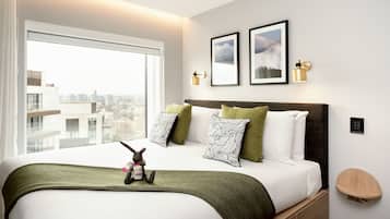 Une chambre d’hôtel moderne avec un grand lit, une vue sur la ville, des œuvres d’art encadrées et une petite table de chevet.